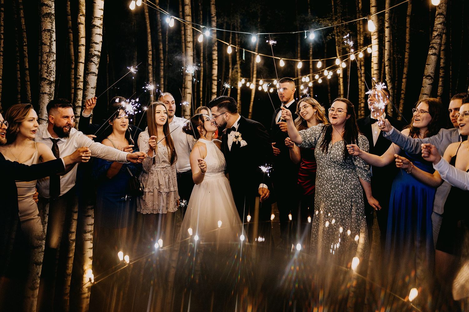 Wedding couple kissing surrounded by guests holding sparklers under string lights in a forest setting.