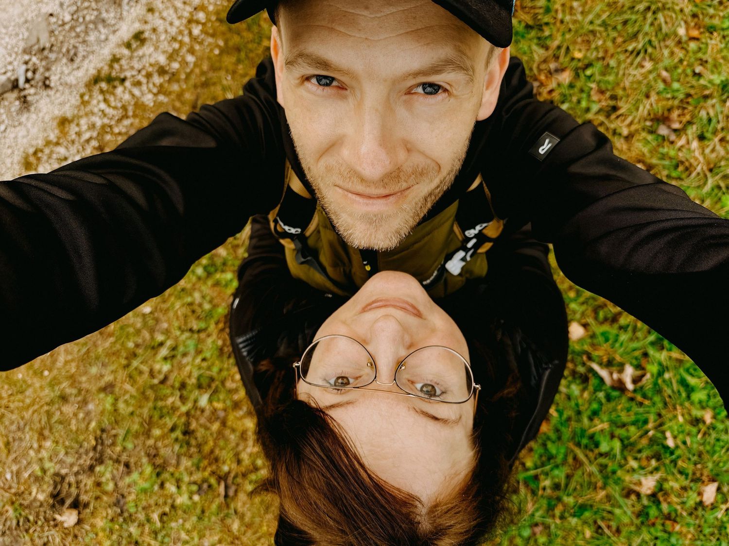 Two people take a fun selfie outdoors, one upside down wearing glasses, both smiling near green grass.