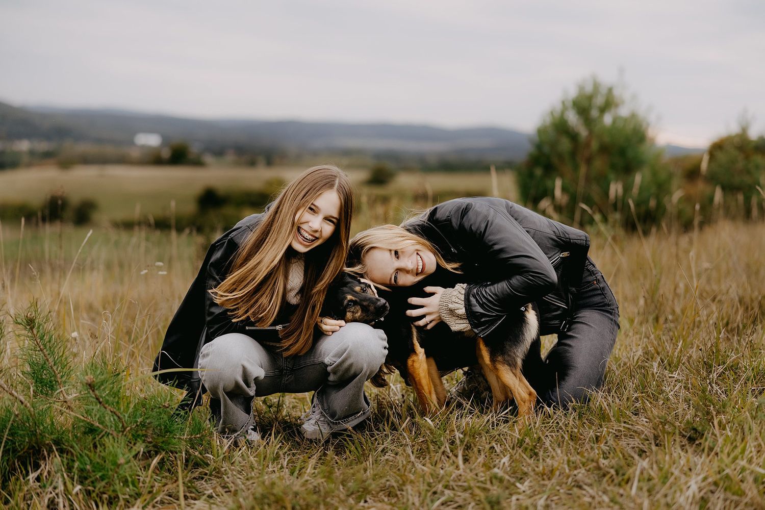 Two teenage girls smiling and playing with a small dog in an open grassy field.