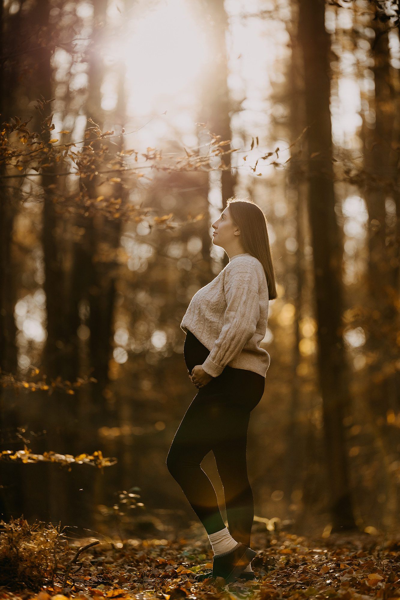 Pregnant woman in cream sweater standing in golden autumn forest holding her baby bump.