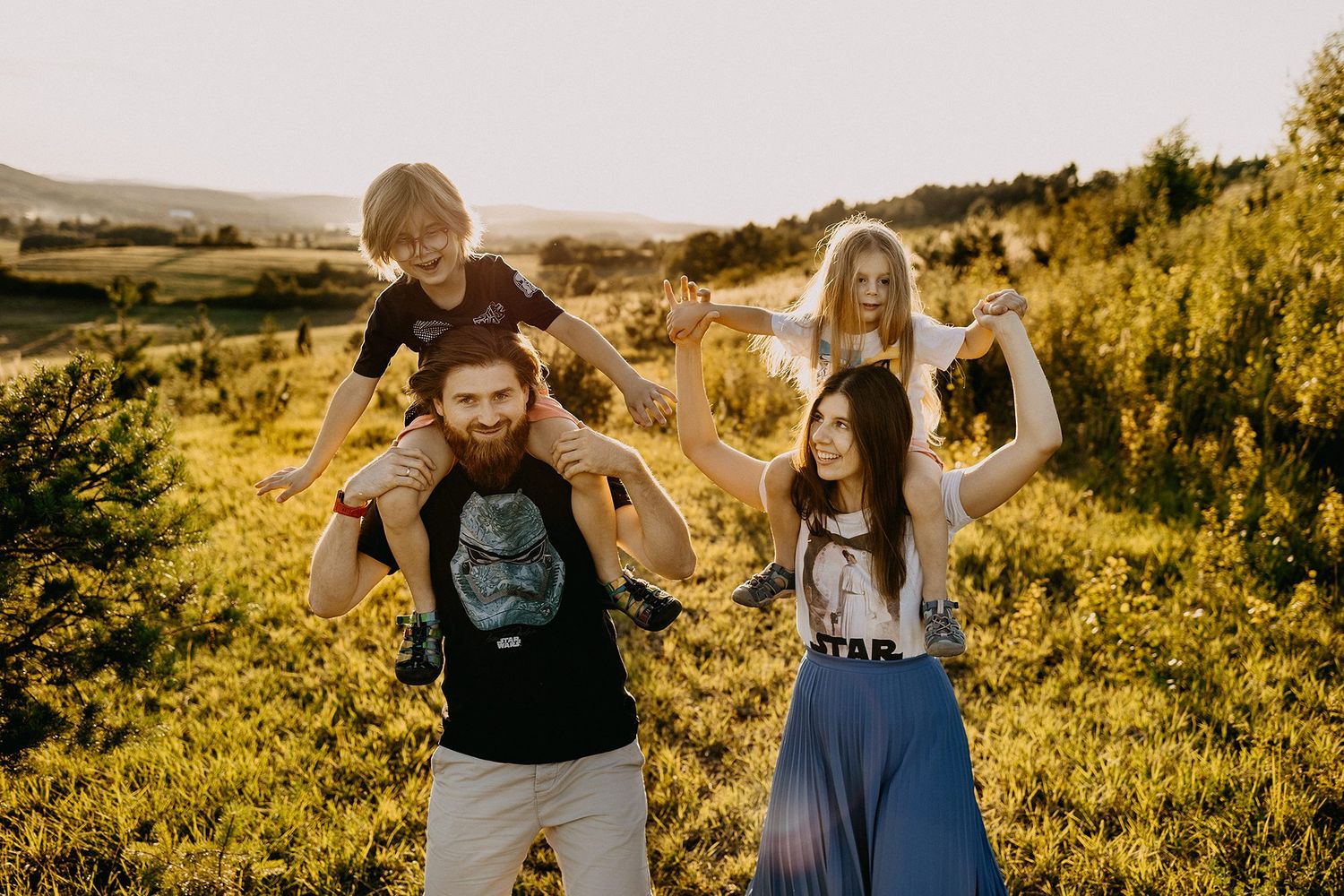 Playful family with two children on parents shoulders enjoying golden sunset in lush green landscape.