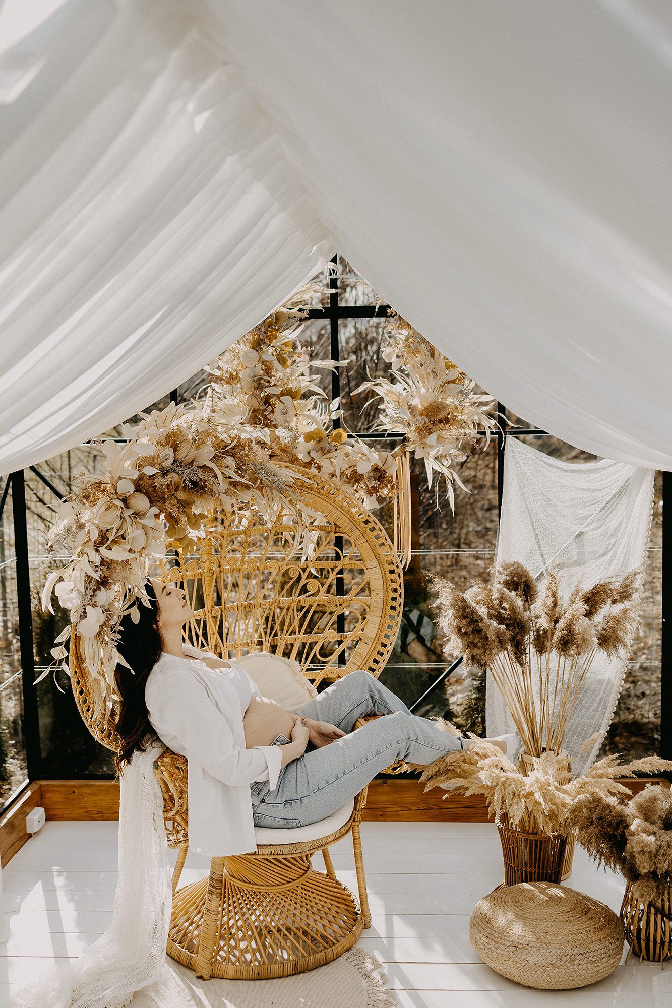 Pregnant woman relaxing in wicker peacock chair surrounded by dried floral arrangements in boho studio.