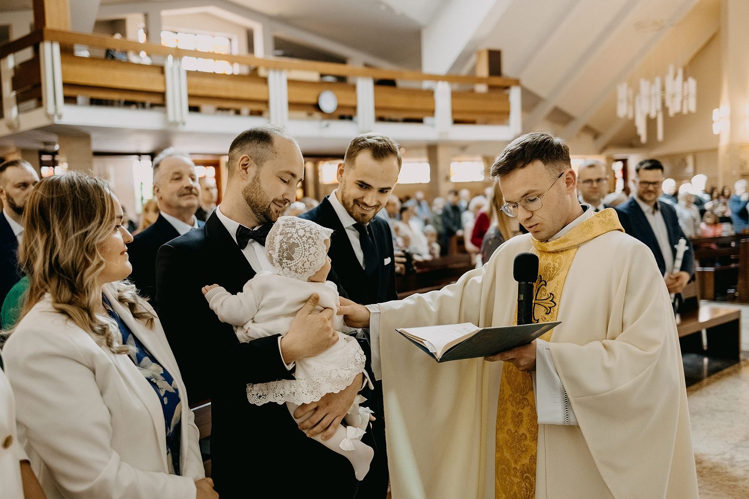 Priest reads from book during Catholic baptism ceremony with godfather holding infant in white christening gown.