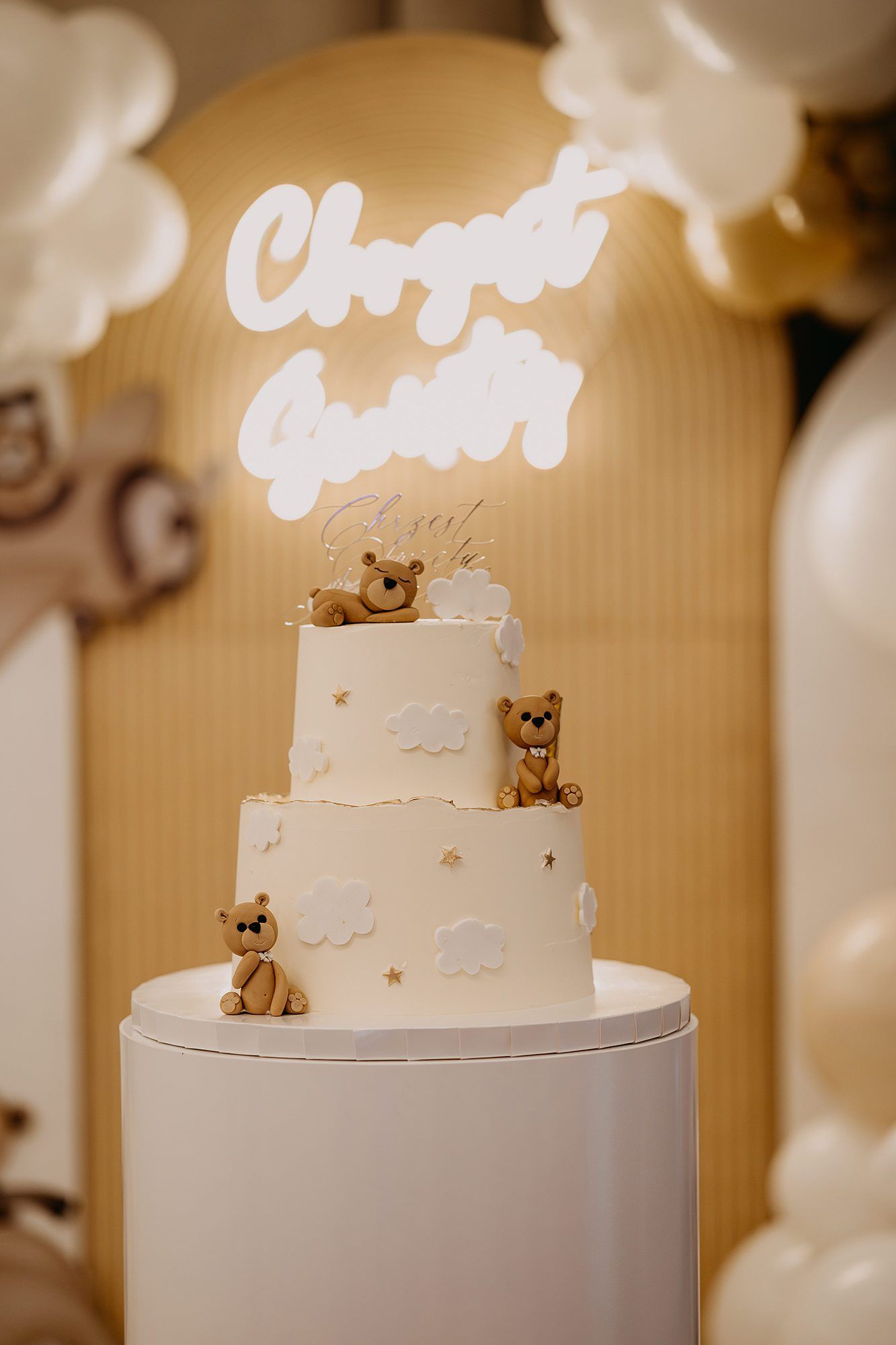 Elegant three-tier white christening cake decorated with teddy bears under glowing neon sign.