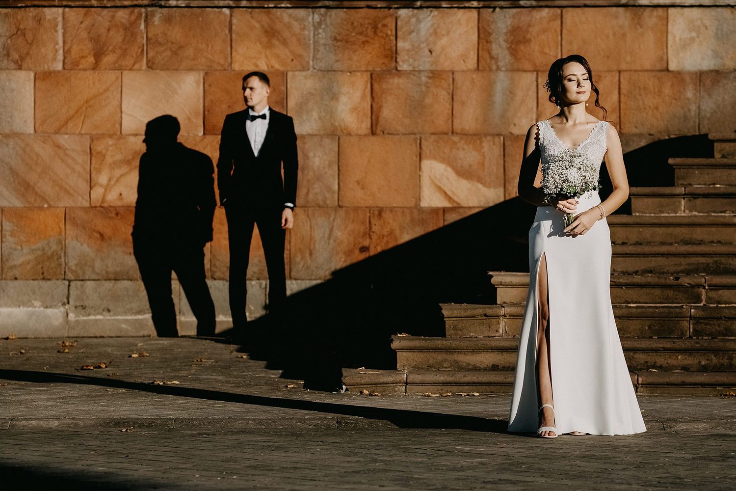 Elegant bride in slit gown with groom casting dramatic shadows on stone steps at sunset.