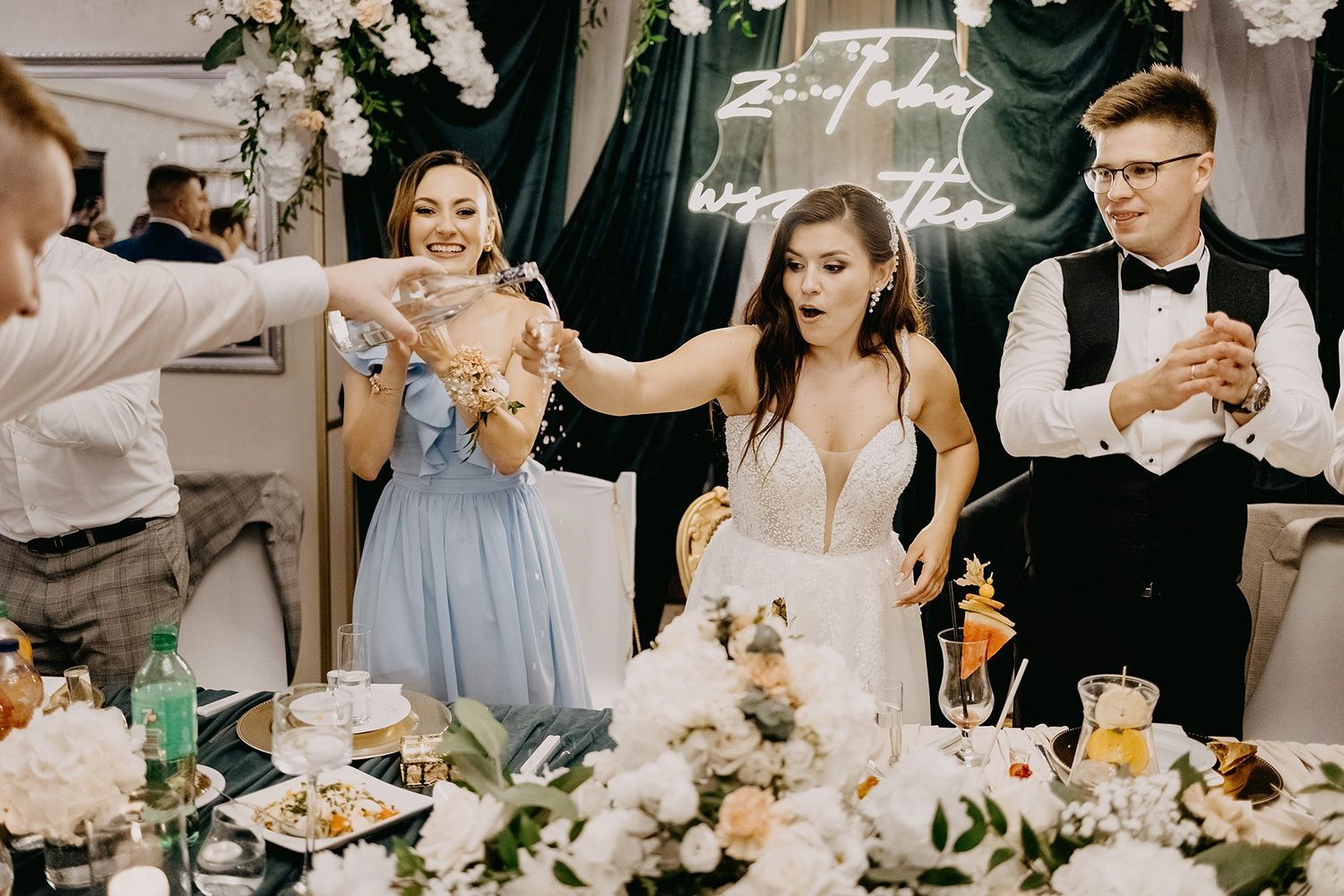 Bride in plunging white gown celebrates with guests at elegantly decorated wedding reception table with white flowers.
