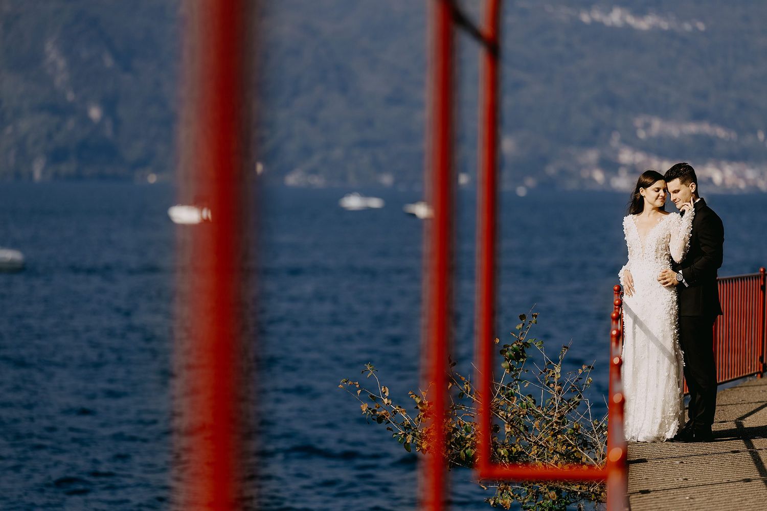 Newlyweds embrace on waterfront dock framed by red railings with stunning mountain lake backdrop.
