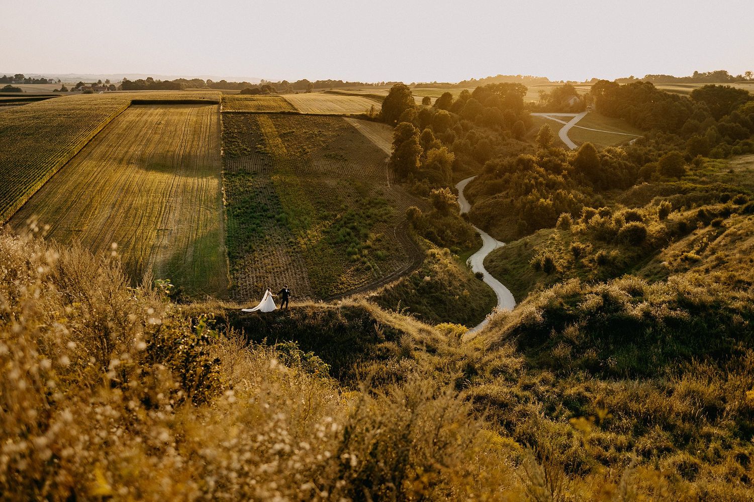 Bride and groom stand on a hillside overlooking a golden sunset countryside landscape.