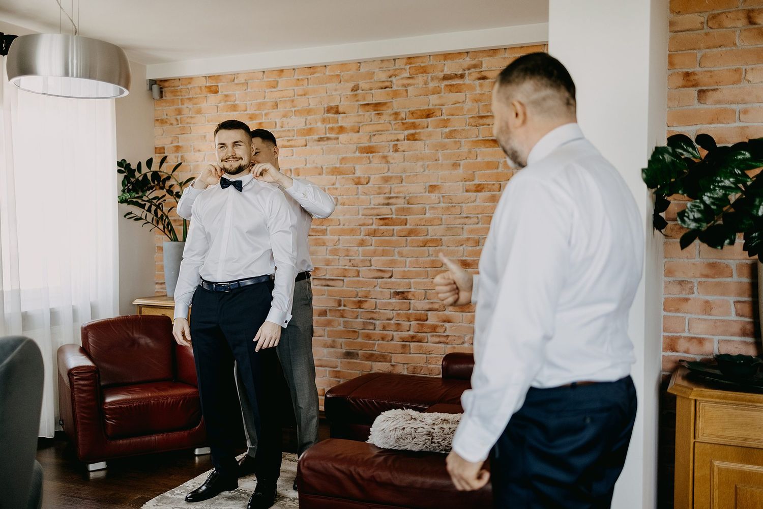 Groomsmen help each other get dressed and adjust bow ties during wedding morning preparations at home.
