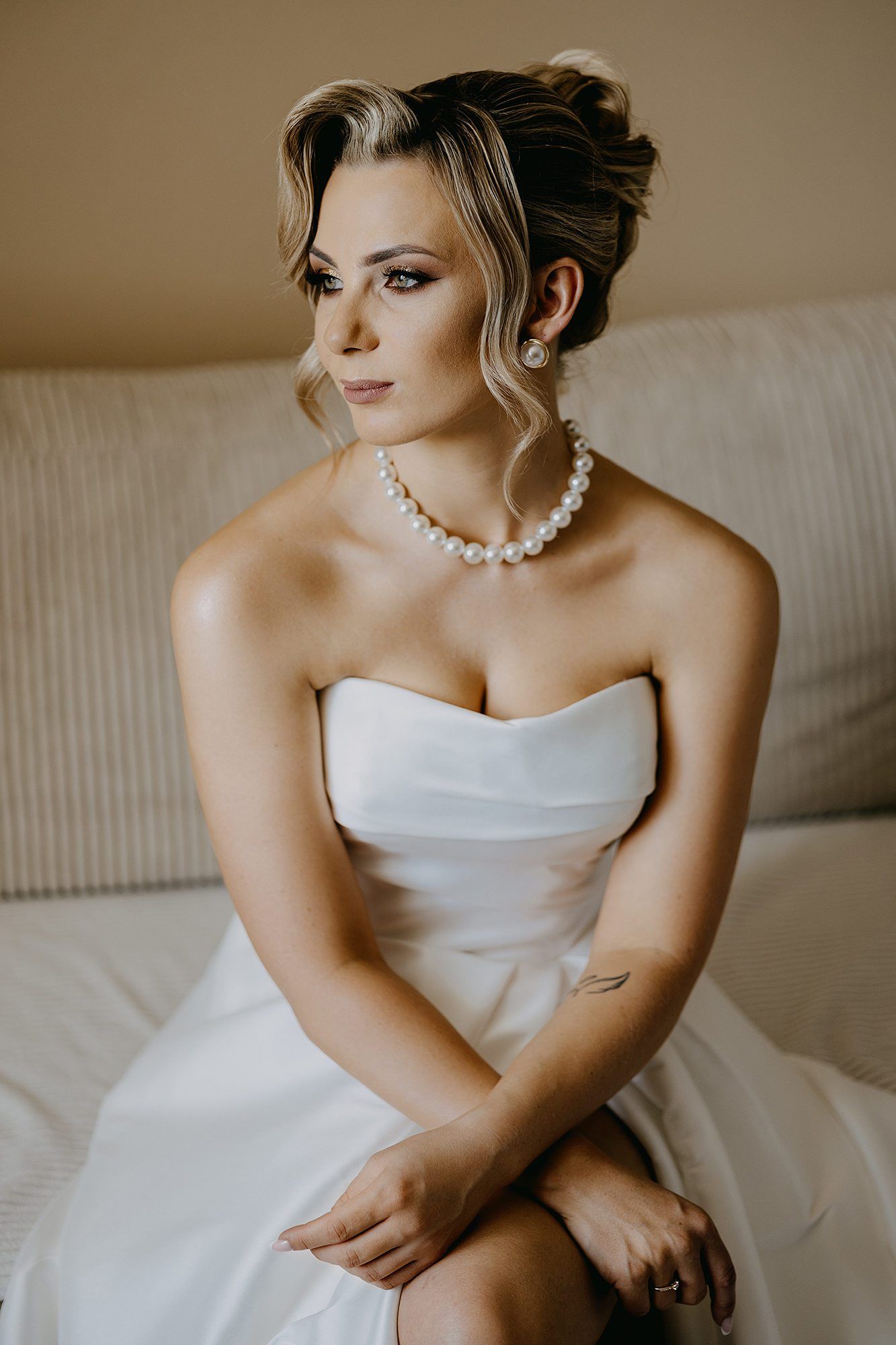 Elegant bride in strapless white gown wearing pearl necklace and earrings, seated on a beige sofa.