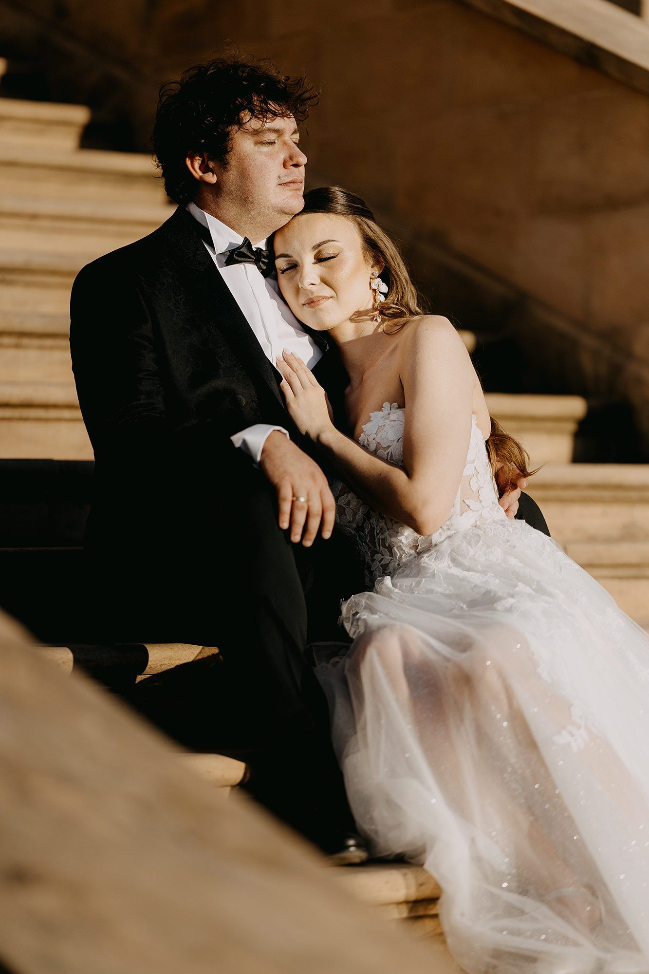 Bride and groom sitting peacefully on stone steps in warm sunlight, eyes closed, embracing.