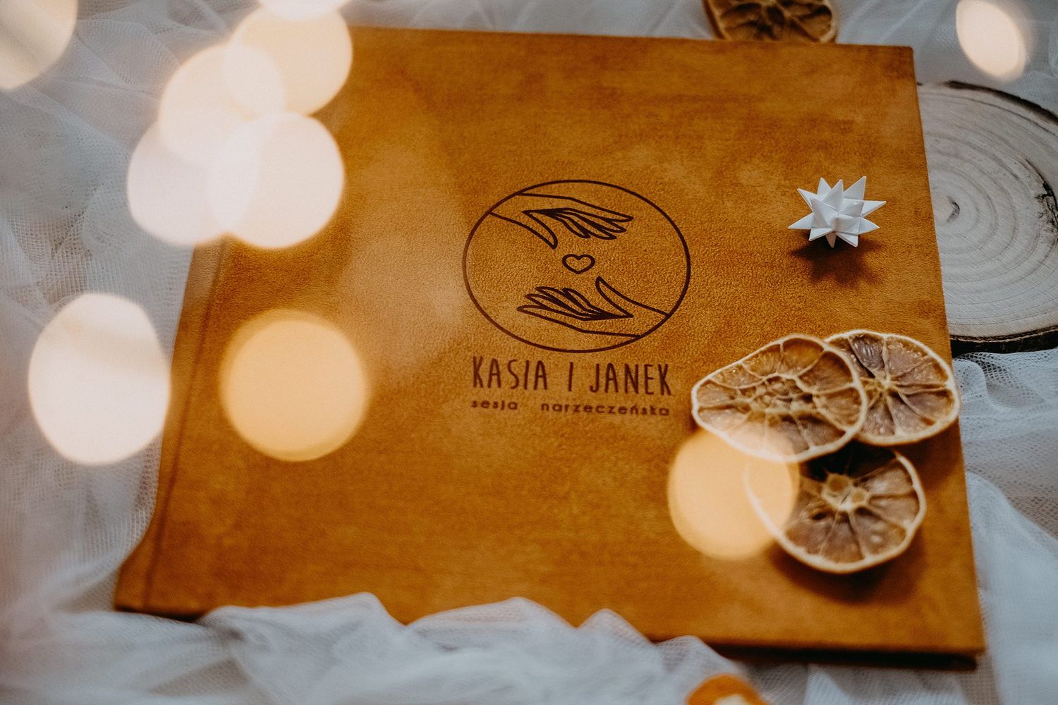 Brown leather wedding photo album cover with circular logo design reading Kasia i Janek on white fabric with dried orange slices.