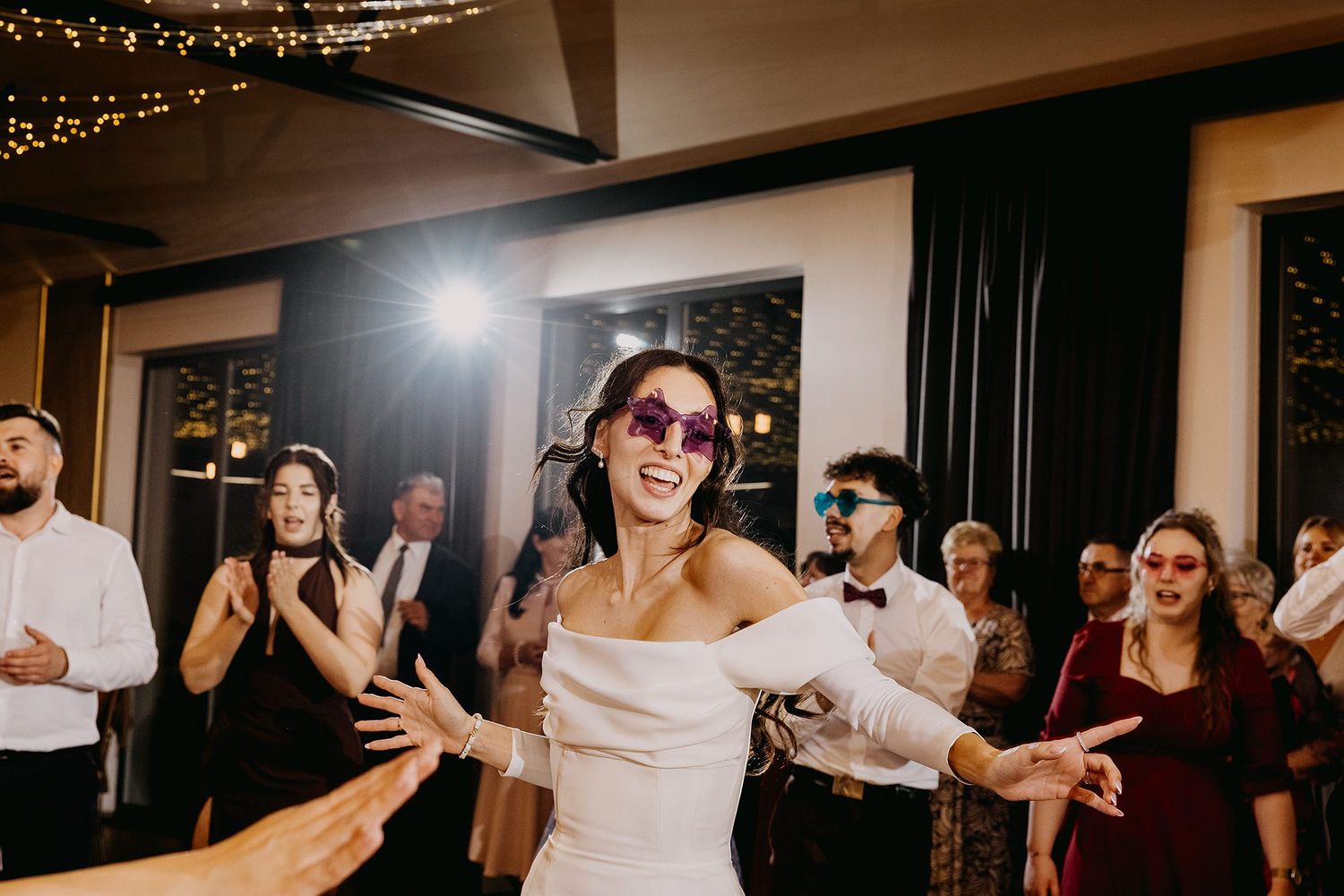 Bride in white off-shoulder gown wearing purple sunglasses dancing joyfully with guests at a lively wedding reception.