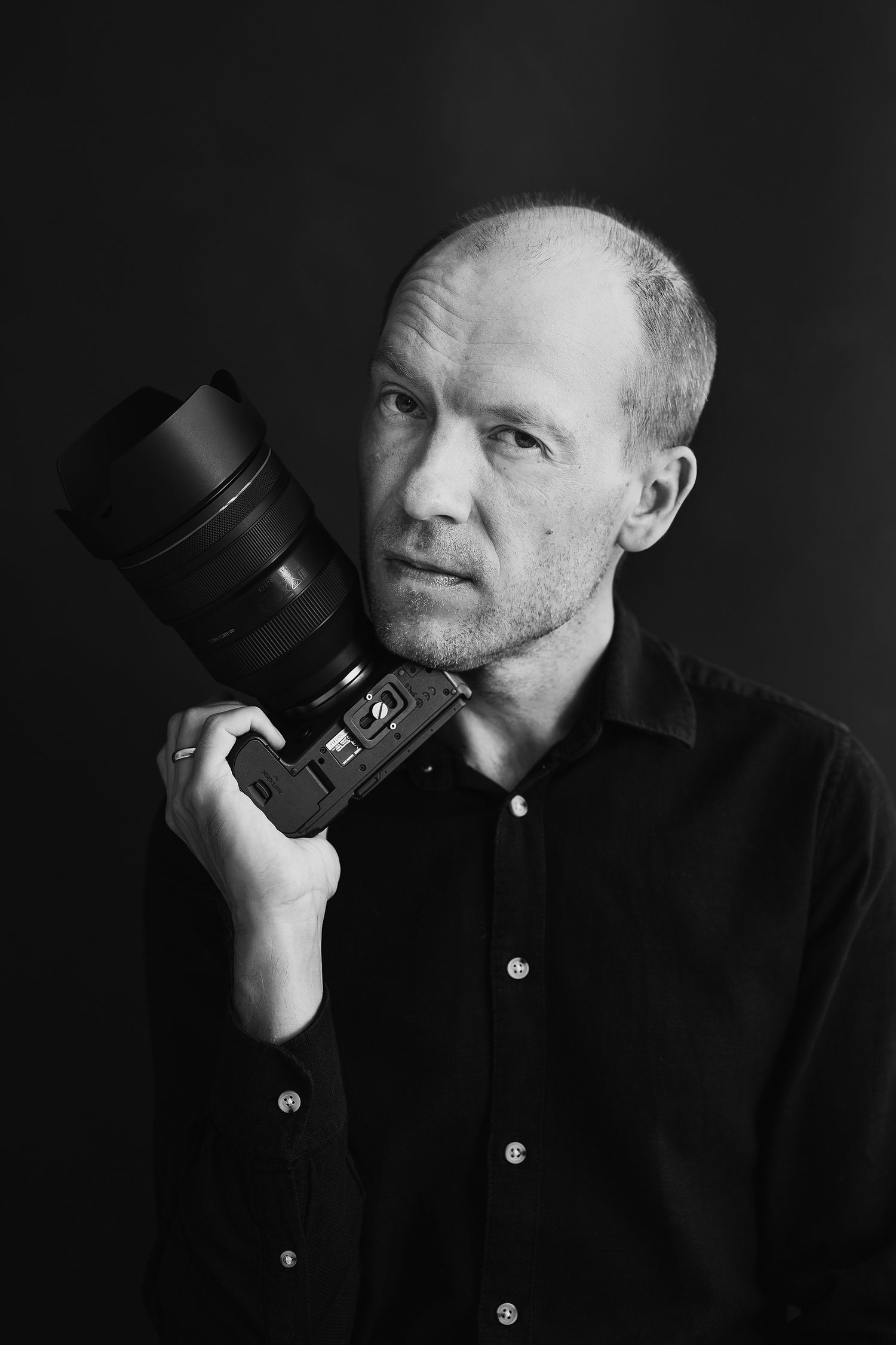 Black and white portrait of a bald man in dark shirt holding a professional DSLR camera with large lens.
