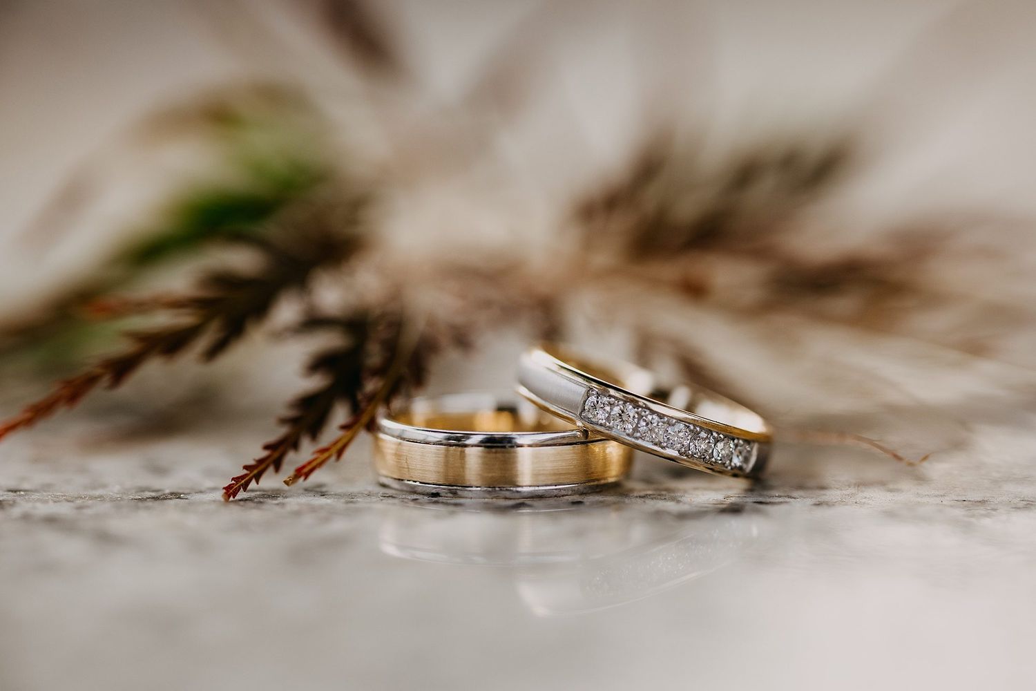 Two elegant wedding bands, one gold and one diamond-encrusted, resting on a marble surface with dried botanicals.