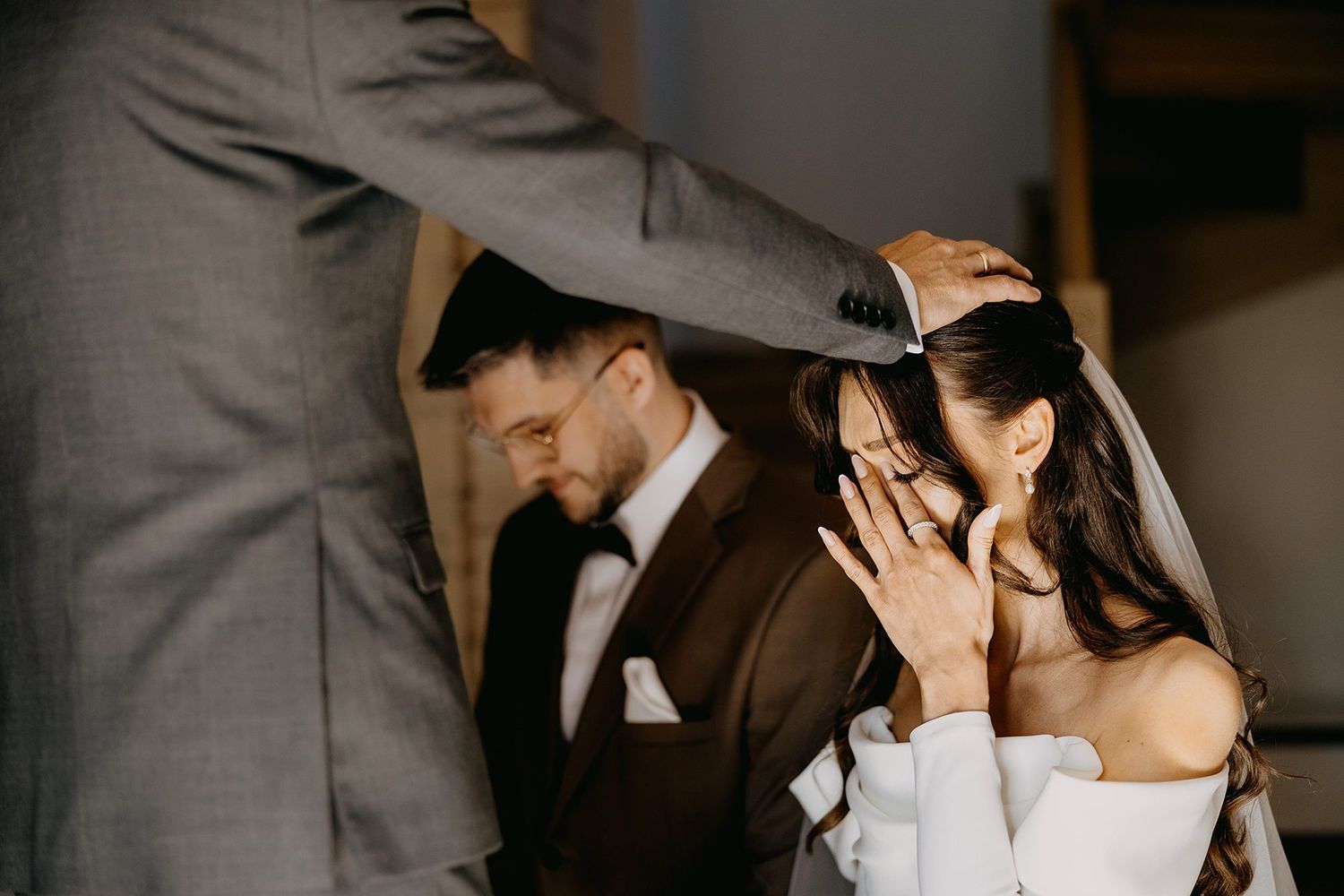 Bride and groom share emotional wedding moment as officiant places veil, bride covering face with hand.
