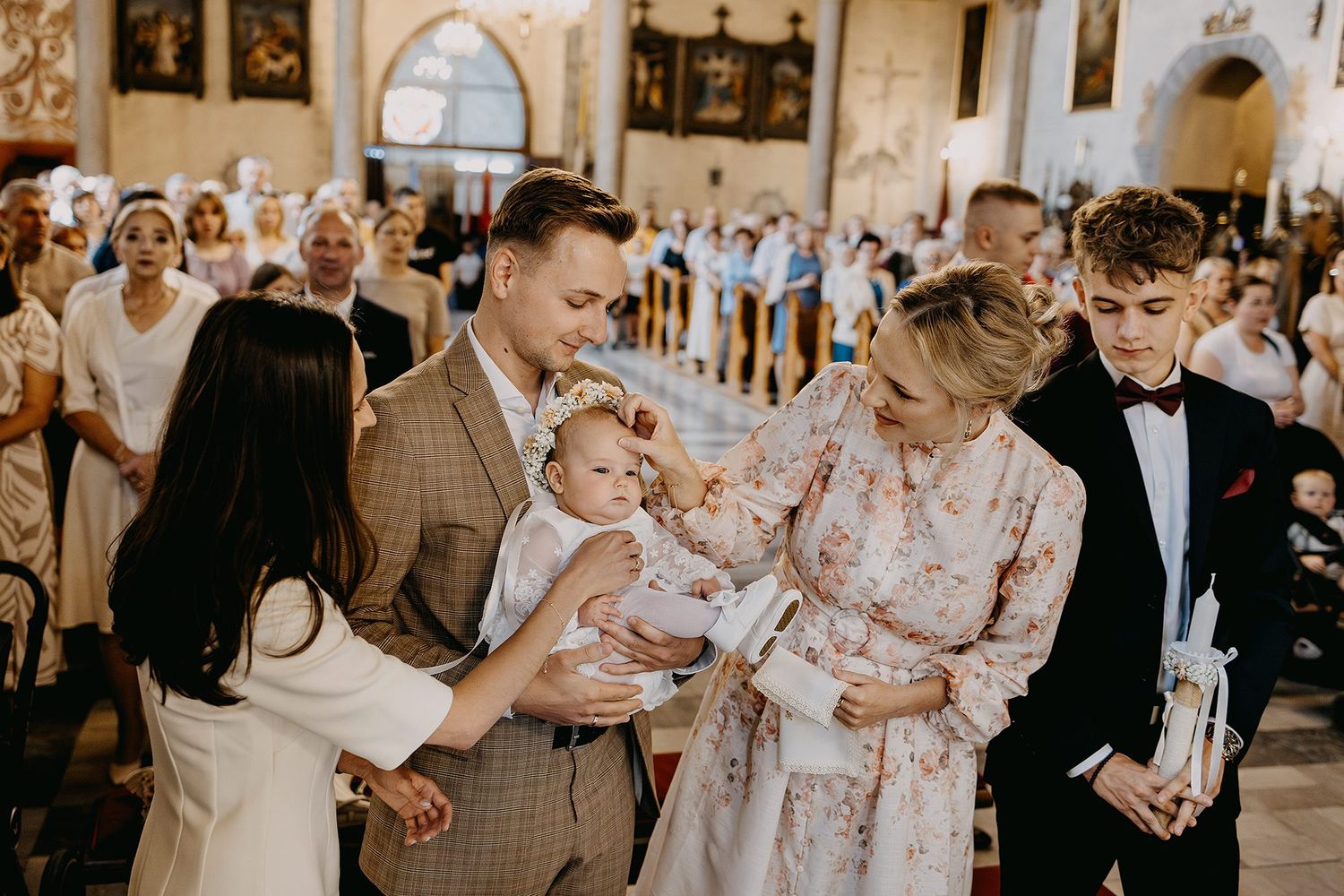 Family and godparents gathered around baby in white christening gown during Catholic baptism church ceremony.