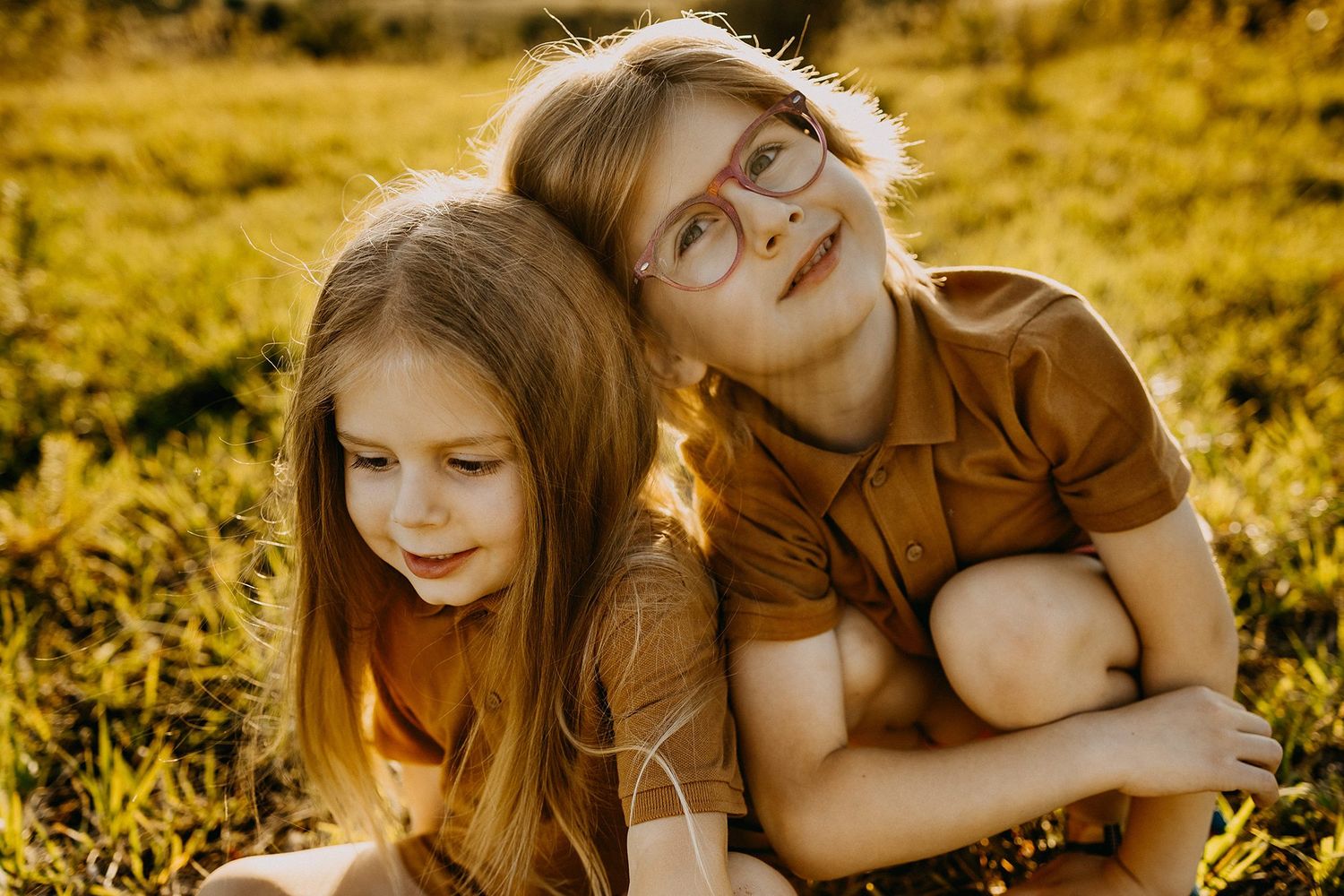 Two young sisters hugging and smiling in sunlit grass, one wearing glasses and a brown shirt.