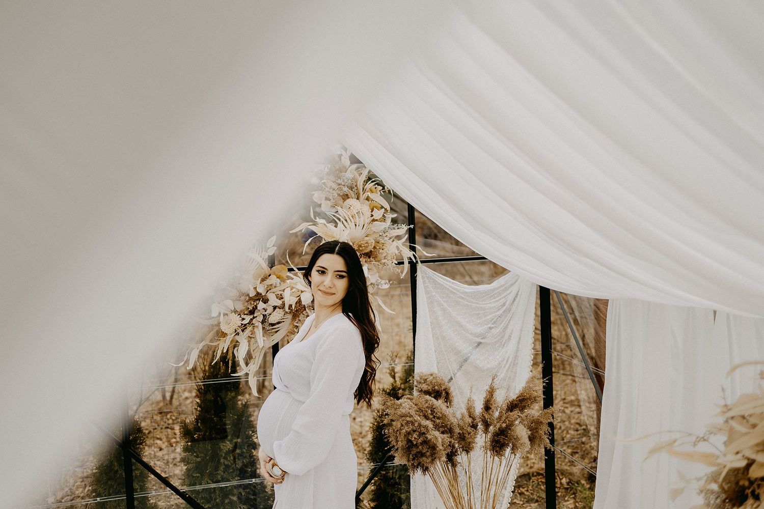 Pregnant woman in white dress posing gracefully among boho dried floral arrangements and draping.