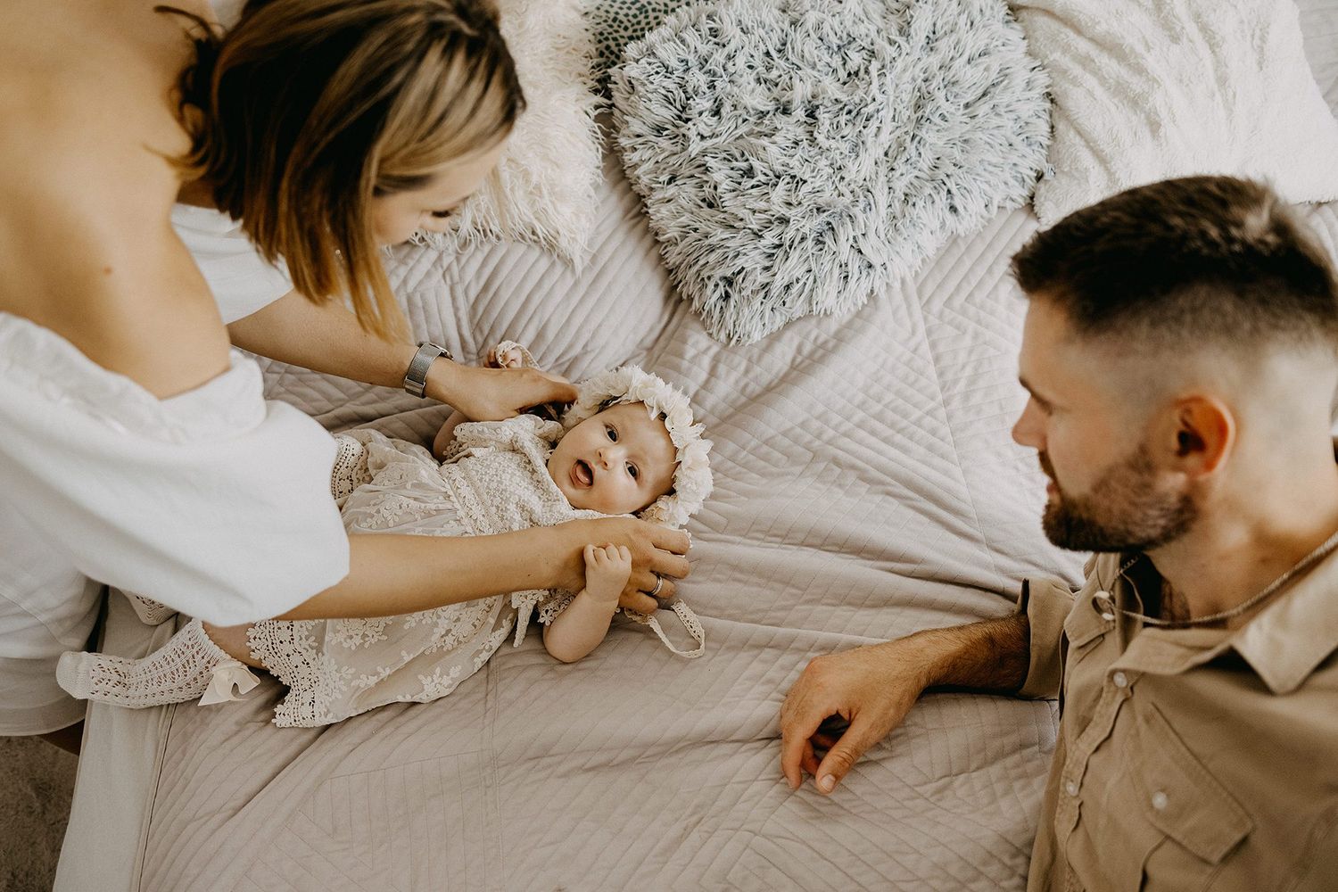 Parents bonding with smiling newborn baby lying on bed during family photo session.