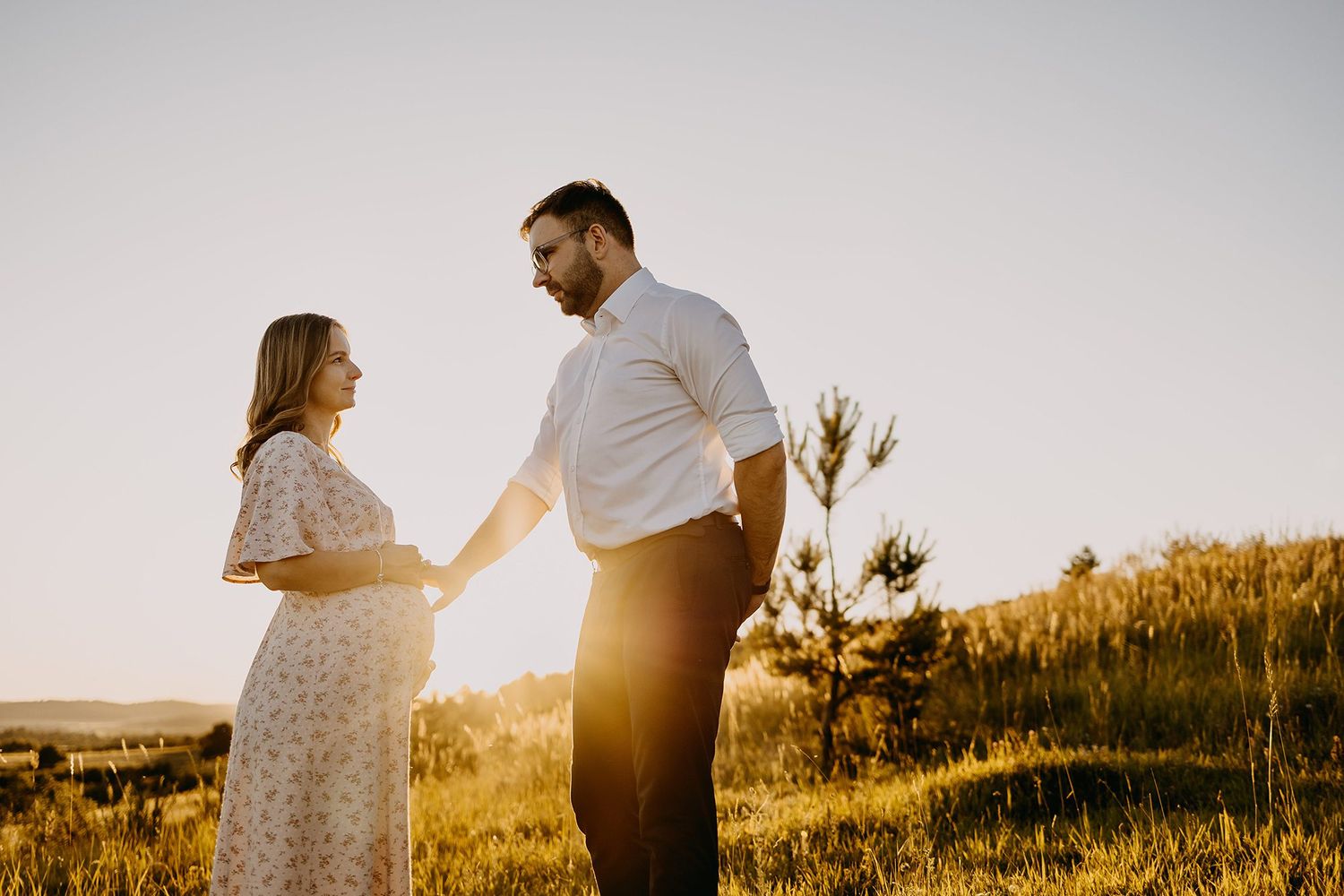 Pregnant couple holding hands facing each other in golden sunset light on grassy hillside.