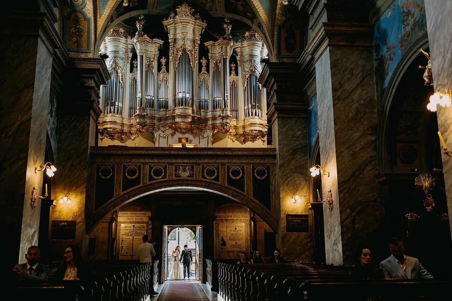 A couple stands silhouetted in a grand church doorway beneath an ornate pipe organ during their wedding.