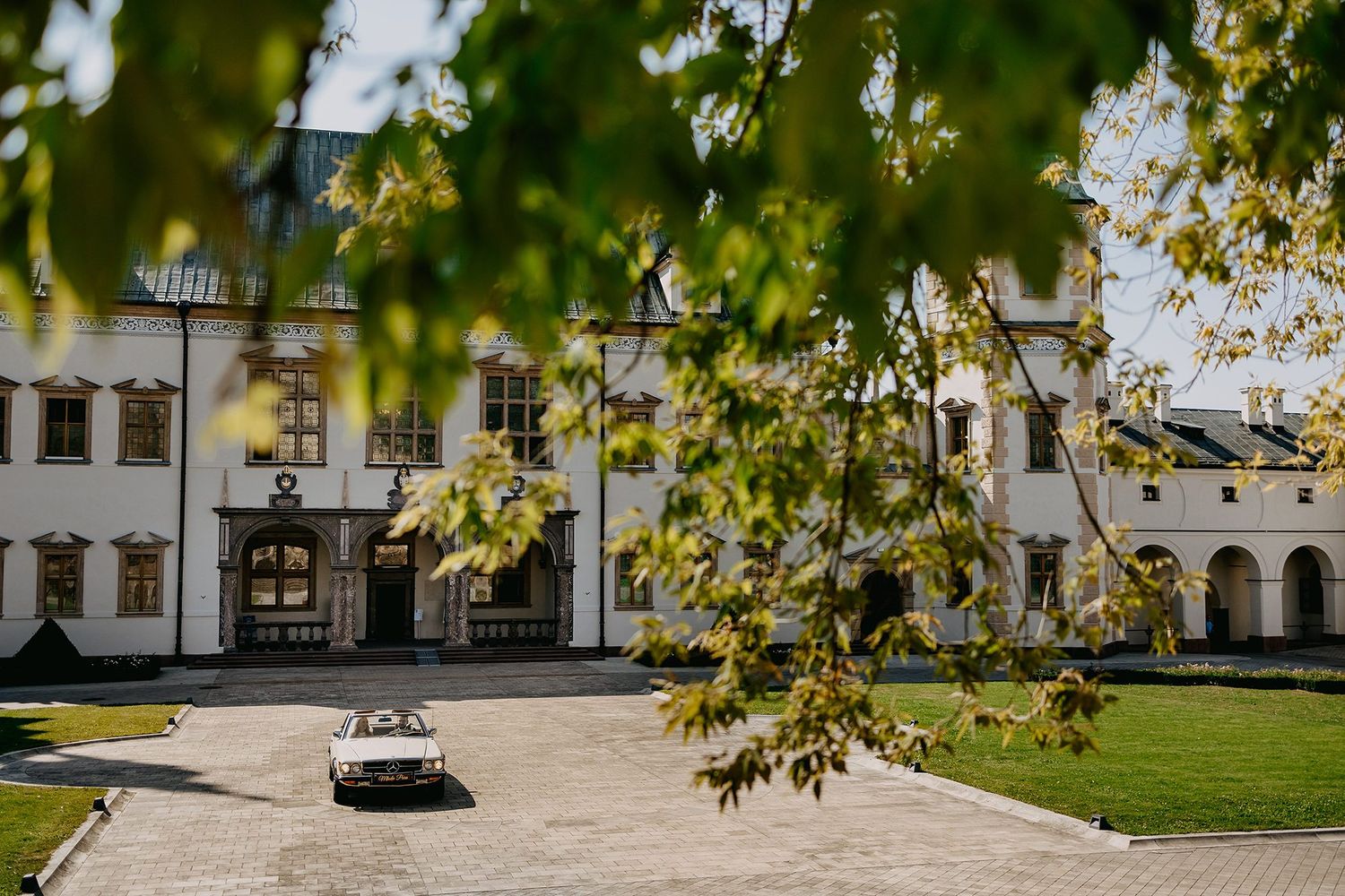 Elegant white historic manor building framed by green leaves with vintage car in courtyard.