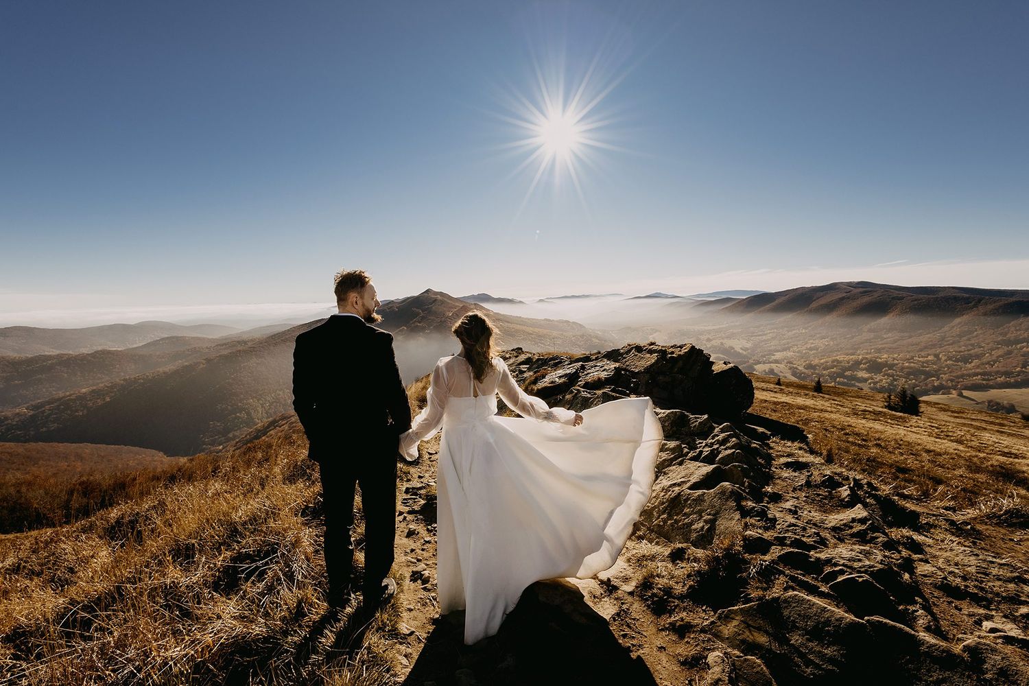 Wedding couple standing on mountain summit with stunning panoramic valley views under bright sun.