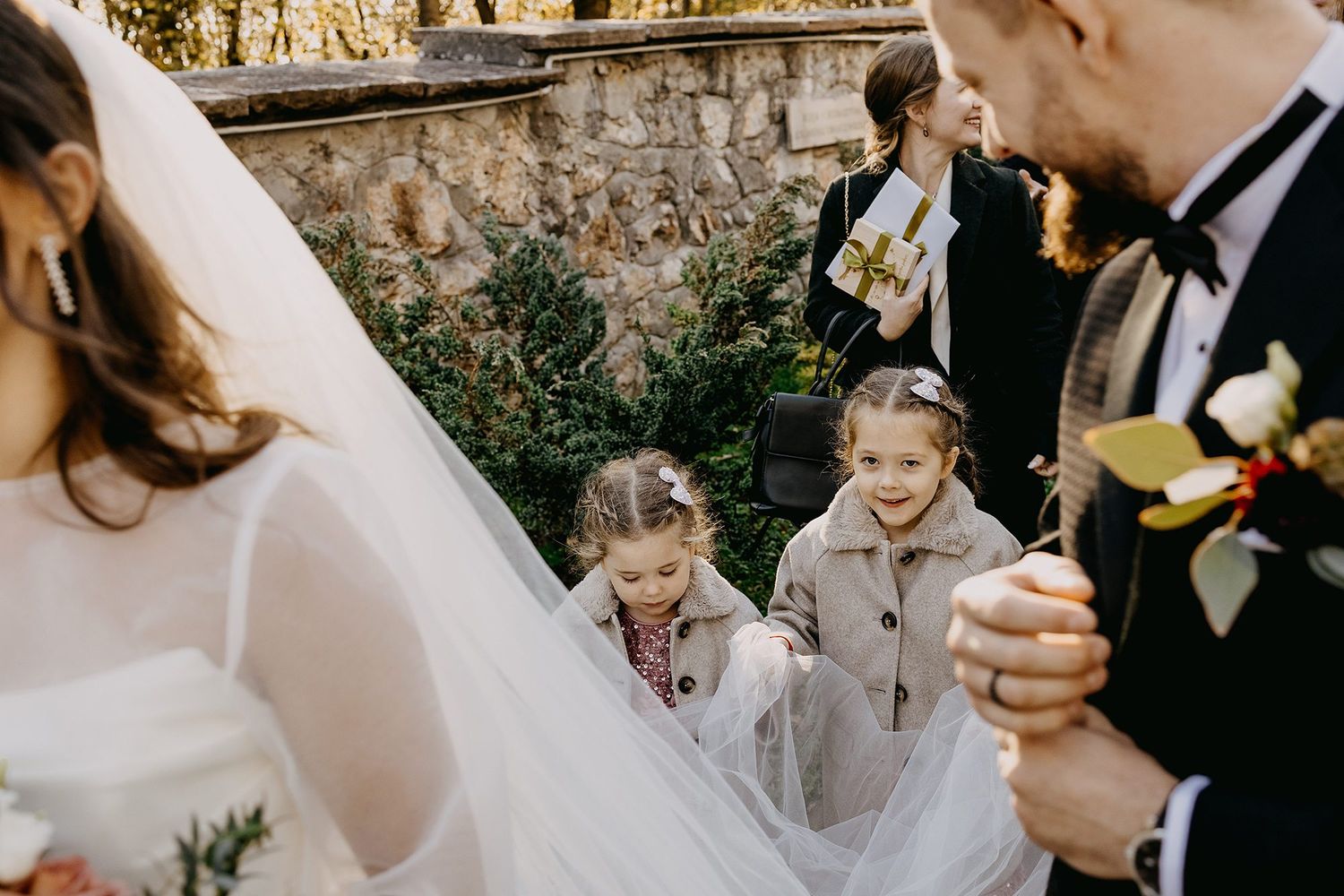 Adorable flower girls playfully lift bride's veil train during outdoor wedding celebration.