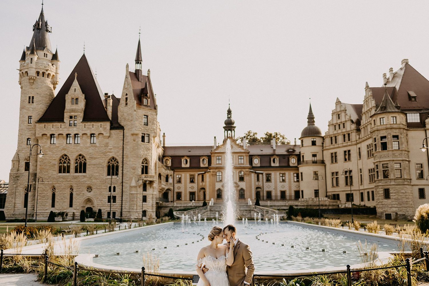 Wedding couple embraces near a grand fountain in front of a stunning Gothic revival castle.
