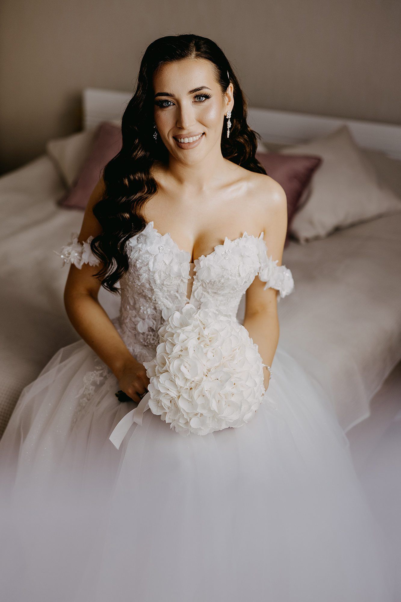 Beautiful brunette bride in off-shoulder white lace gown holding white hydrangea bouquet on bed.
