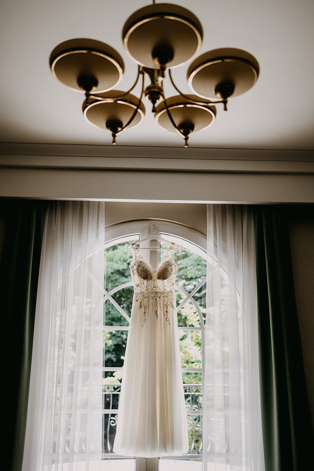 A beautiful lace wedding dress hangs elegantly from a chandelier in front of an arched window with sheer curtains.