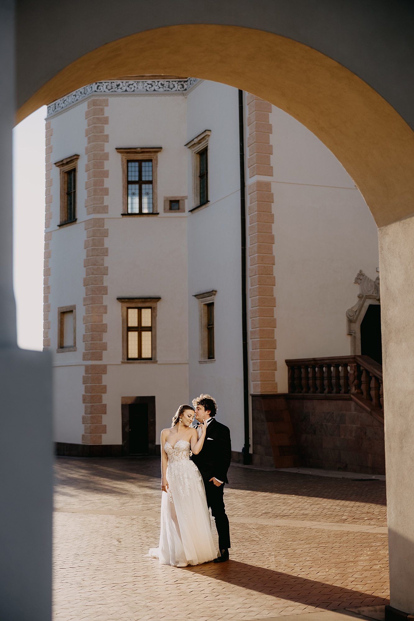 Bride and groom embrace in sunlit courtyard of historic European castle framed by stone archway.