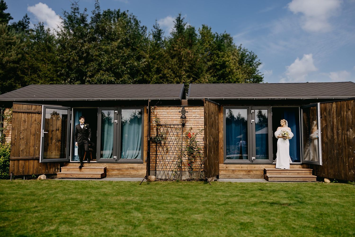 Bride in white gown stands outside modern wooden wedding venue on sunny day.
