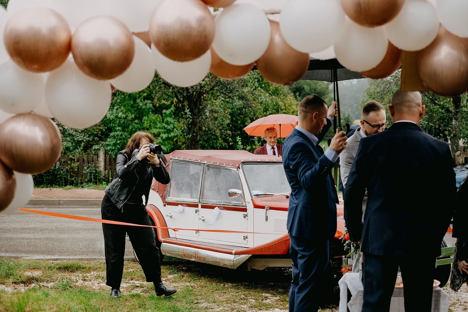 Wedding photographer capturing moments near a vintage orange car under a rose gold balloon arch.