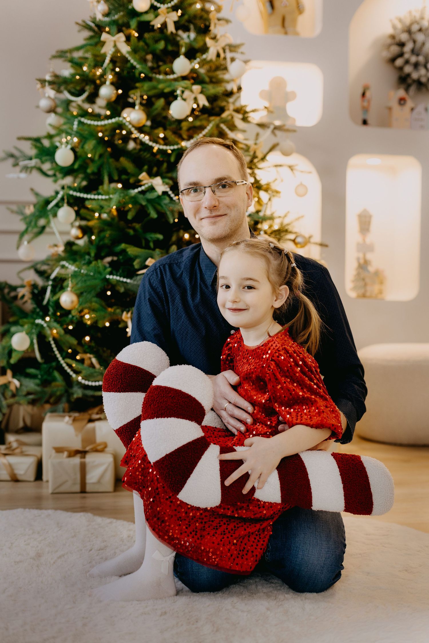 Smiling father and young daughter in a red sequin dress hold a candy cane pillow beside a decorated Christmas tree.