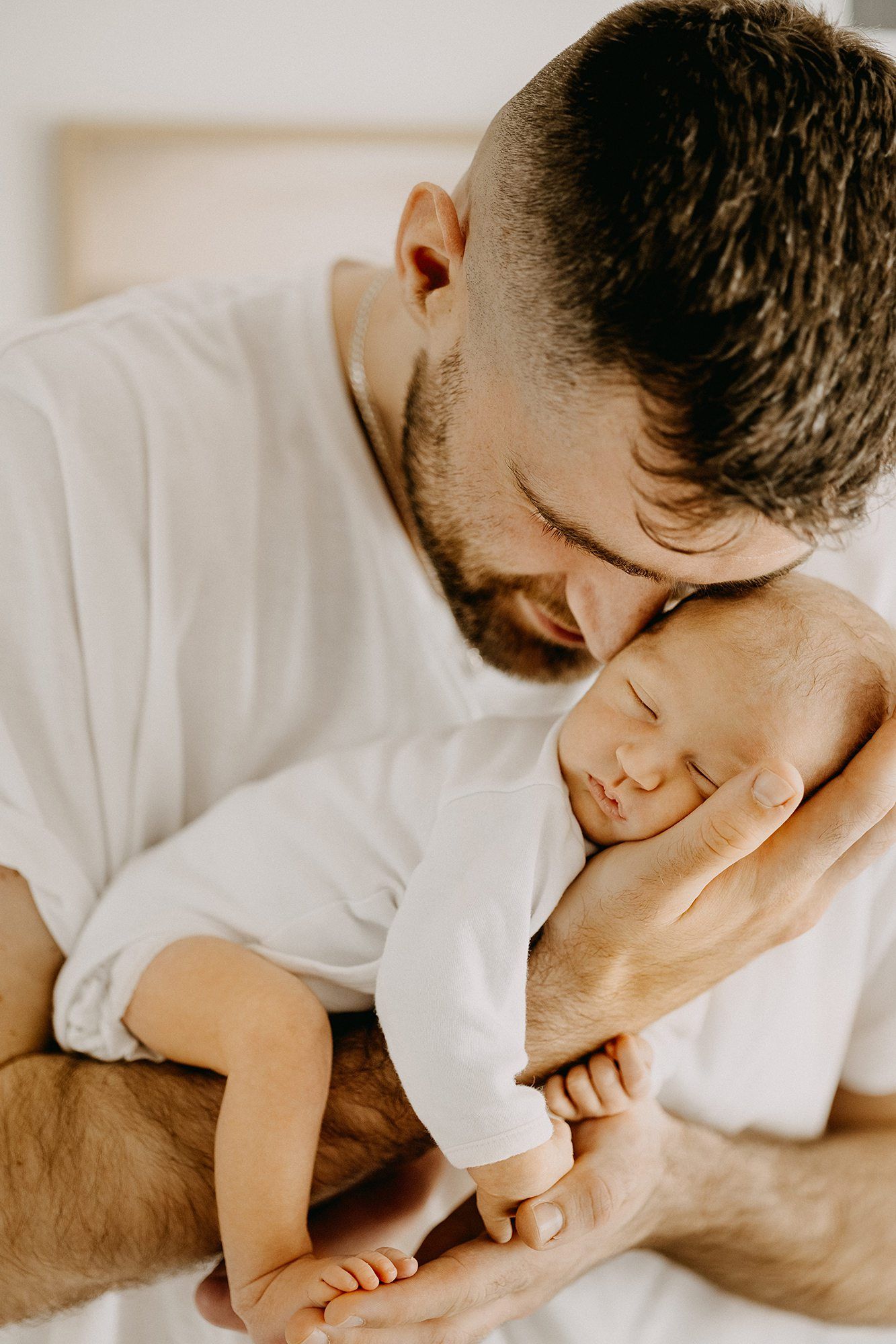 Bearded father in white shirt tenderly kissing sleeping newborn baby cradled in his hands.