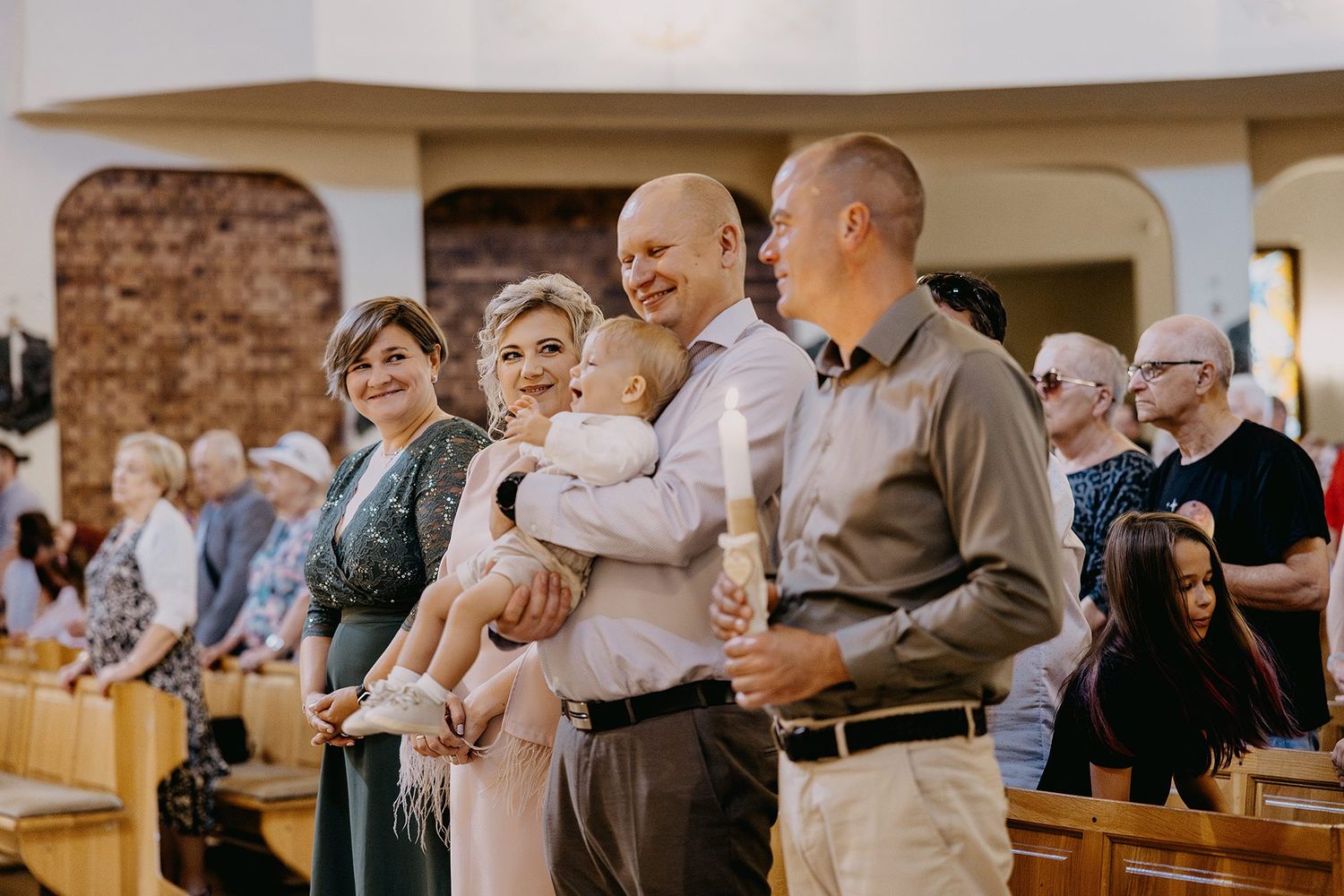 Family standing in church aisle during baptism ceremony with bald father holding baby in white outfit.