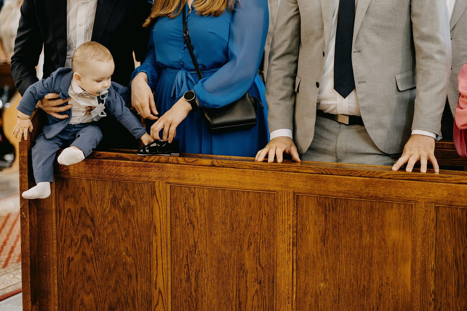 Baby in suit reaches over wooden church pew during baptism ceremony with family.