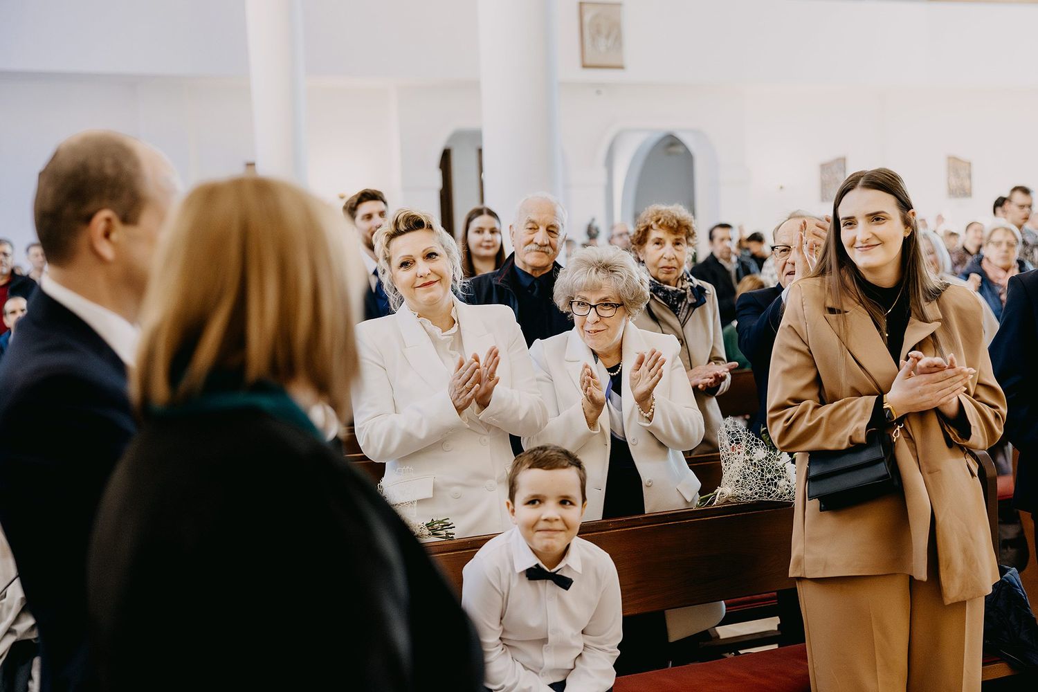 Family and guests applauding during baptism ceremony in church, gathered in wooden pews for celebration.