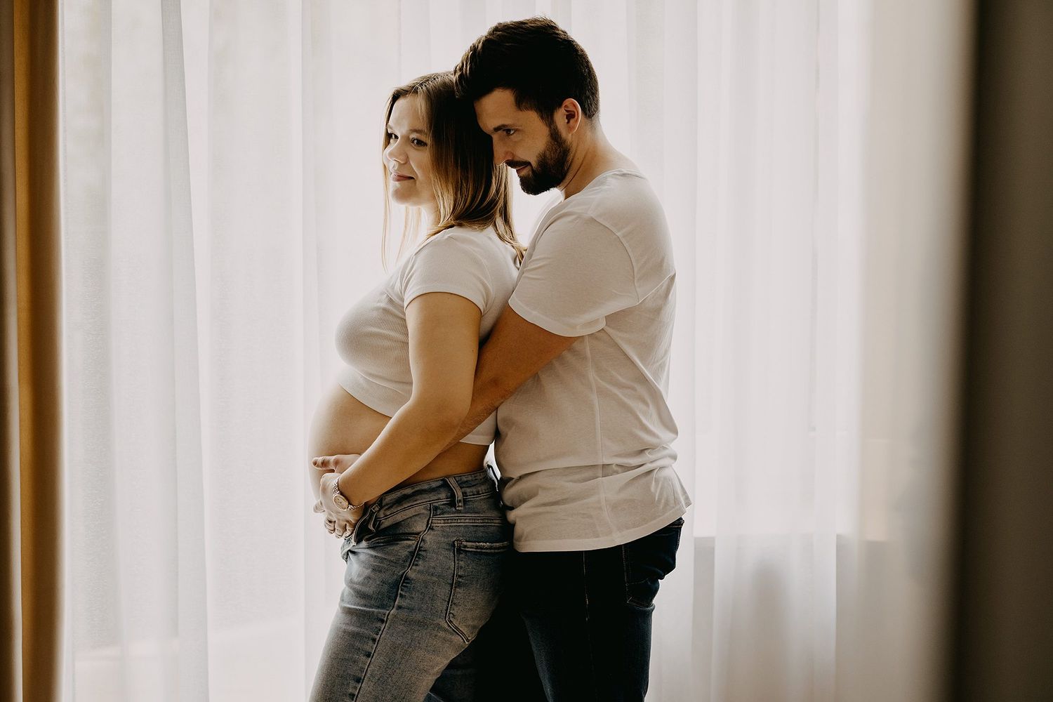 Pregnant couple in white tops standing back-to-front by window, man embracing womans baby bump lovingly.