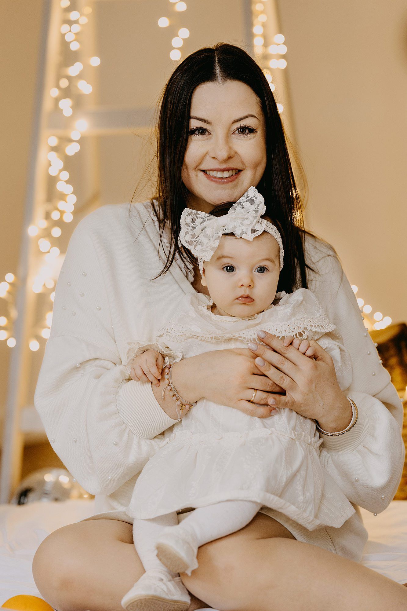 A smiling dark-haired mother in white holds her baby girl with a bow headband before warm bokeh lights.