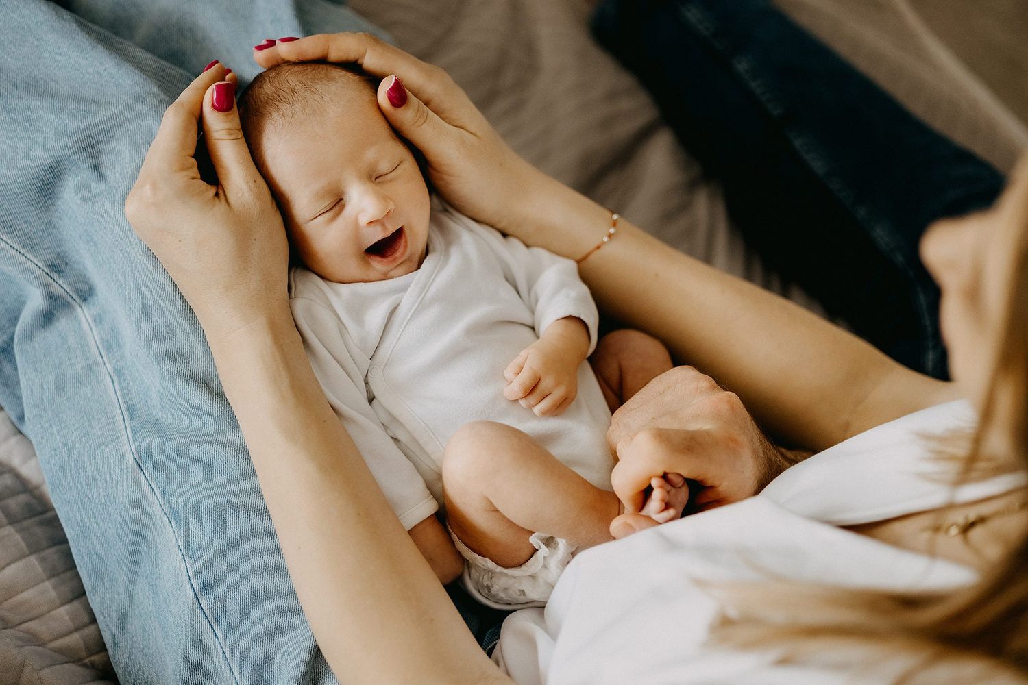 Newborn baby in white onesie yawning while being held in mothers arms on a cozy bed.