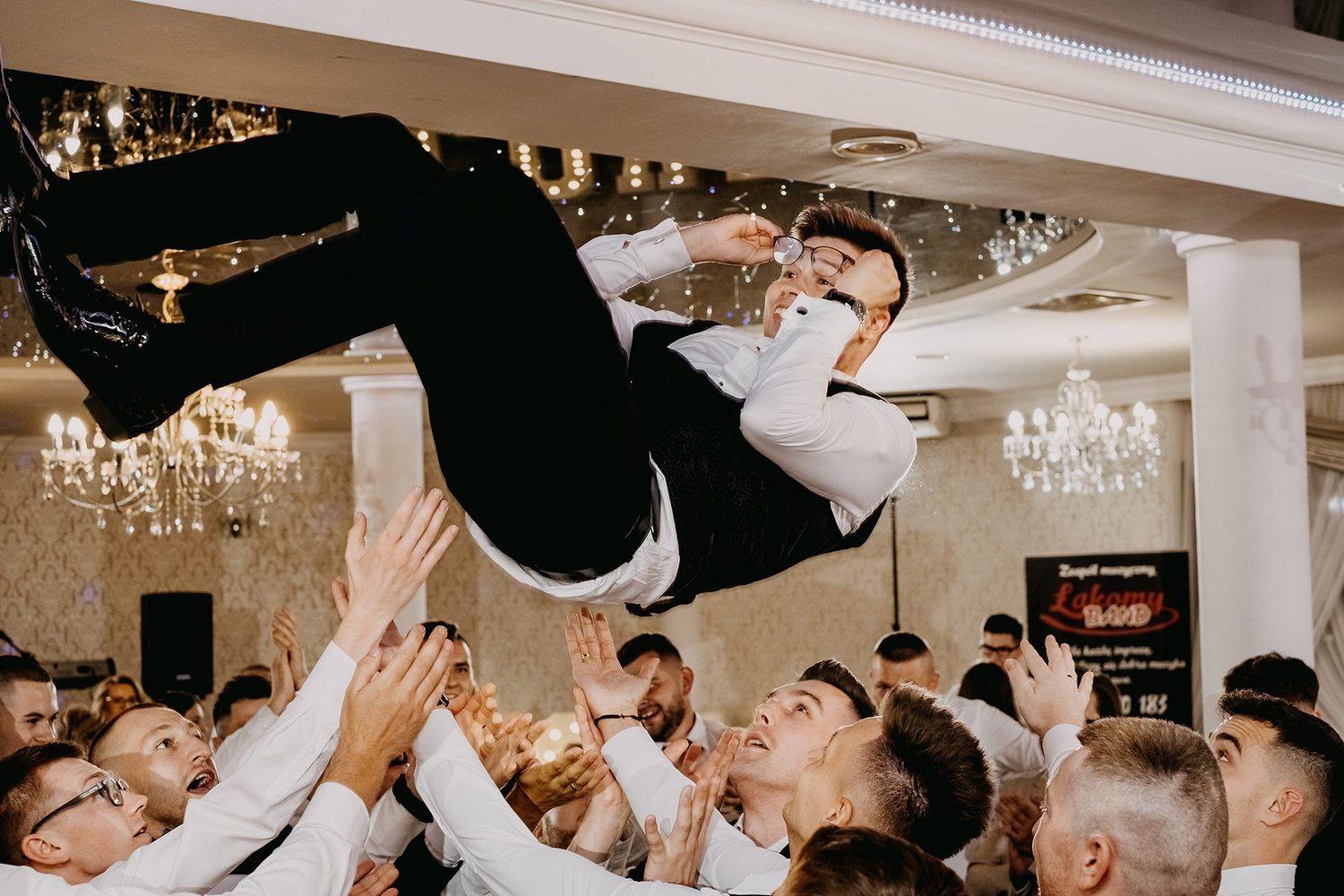 Groom being tossed in the air by groomsmen in white shirts at wedding reception celebration.