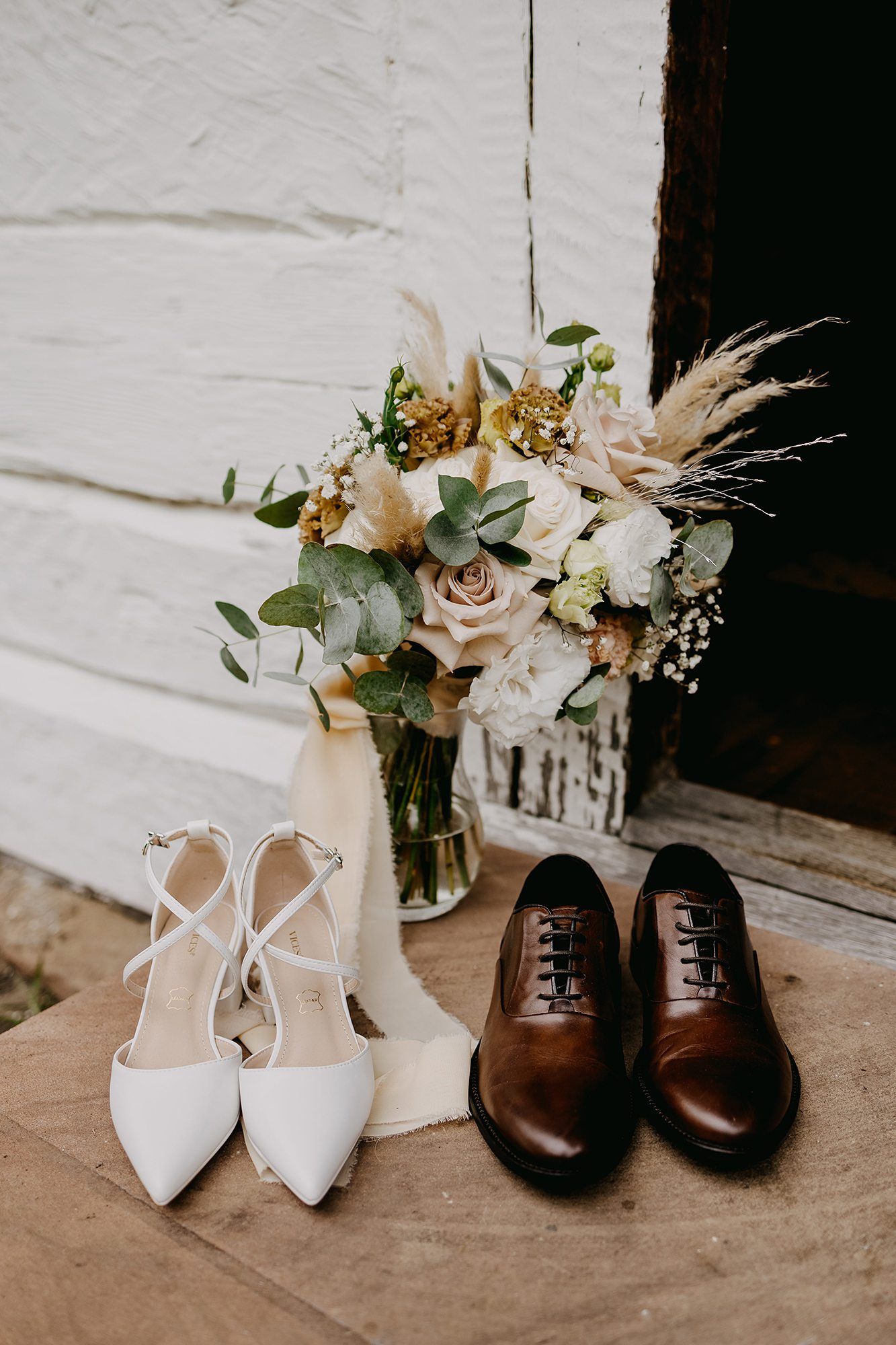 Wedding flat lay featuring white bridal heels, brown leather oxford shoes, and boho floral bouquet.