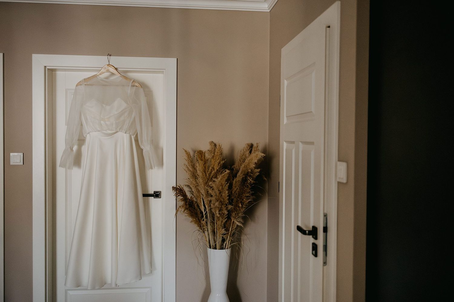 Elegant long-sleeve white wedding dress hanging on a door beside a vase of dried pampas grass.