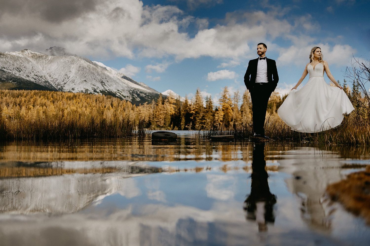 Bride and groom reflected in still water with golden autumn trees and snowy mountains.