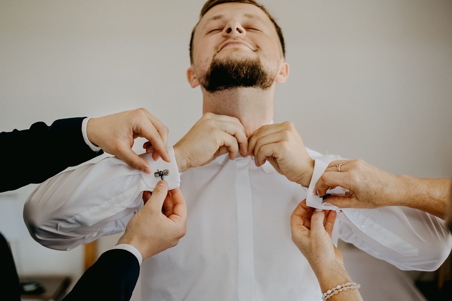 Groomsmen helping groom button his white dress shirt cufflinks during wedding preparation.