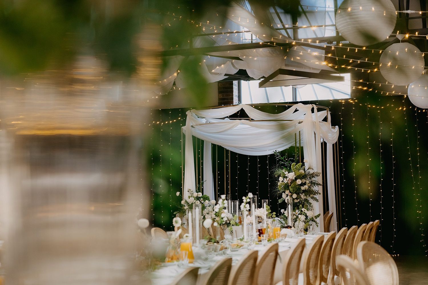 Elegant wedding reception with white draping, string lights, floral centerpieces, and gold chairs in a greenhouse venue.