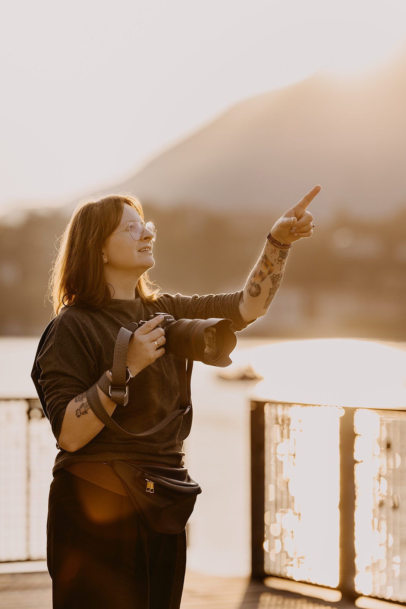 Female photographer with tattoos holding camera pointing at golden hour sunset over mountain lake.